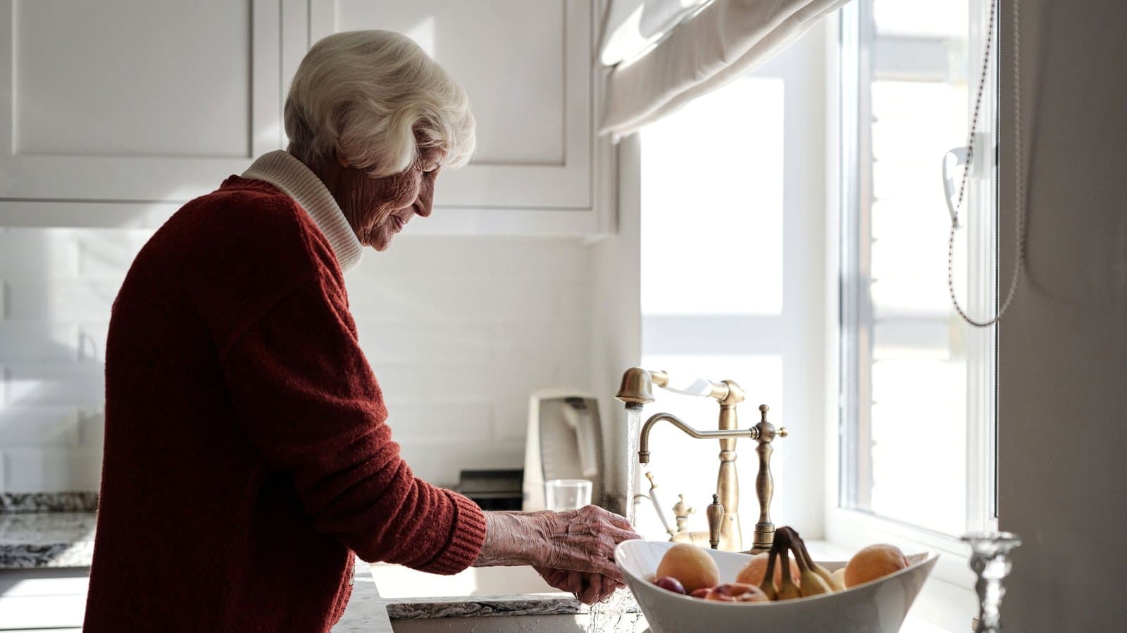 Donna anziana in piedi davanti al lavello della cucina di casa sua, mentre sciacqua la frutta alla luce del giorno che entra dalla finestra